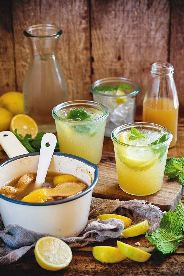 White pot full of ginger syrup and lemons on a wooden table. Three glasses, two with syrup and all with mint and lemon slices. 