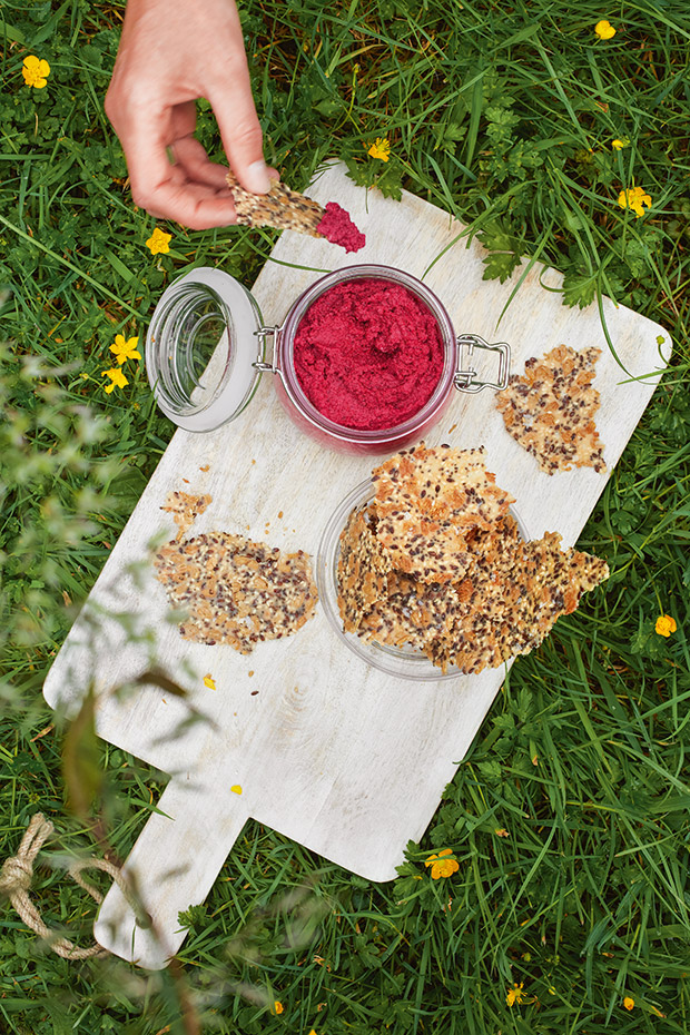 A wooden chopping board with a jar of homemade beetroot cashew dip and a side of seed crackers. 